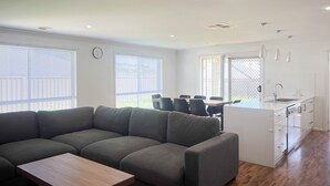 A modern living room with a sectional sofa, a dining table with chairs, and a kitchen area with a sink and cabinets. - Darcy Drive - All of the Comforts (Wagga Wagga)