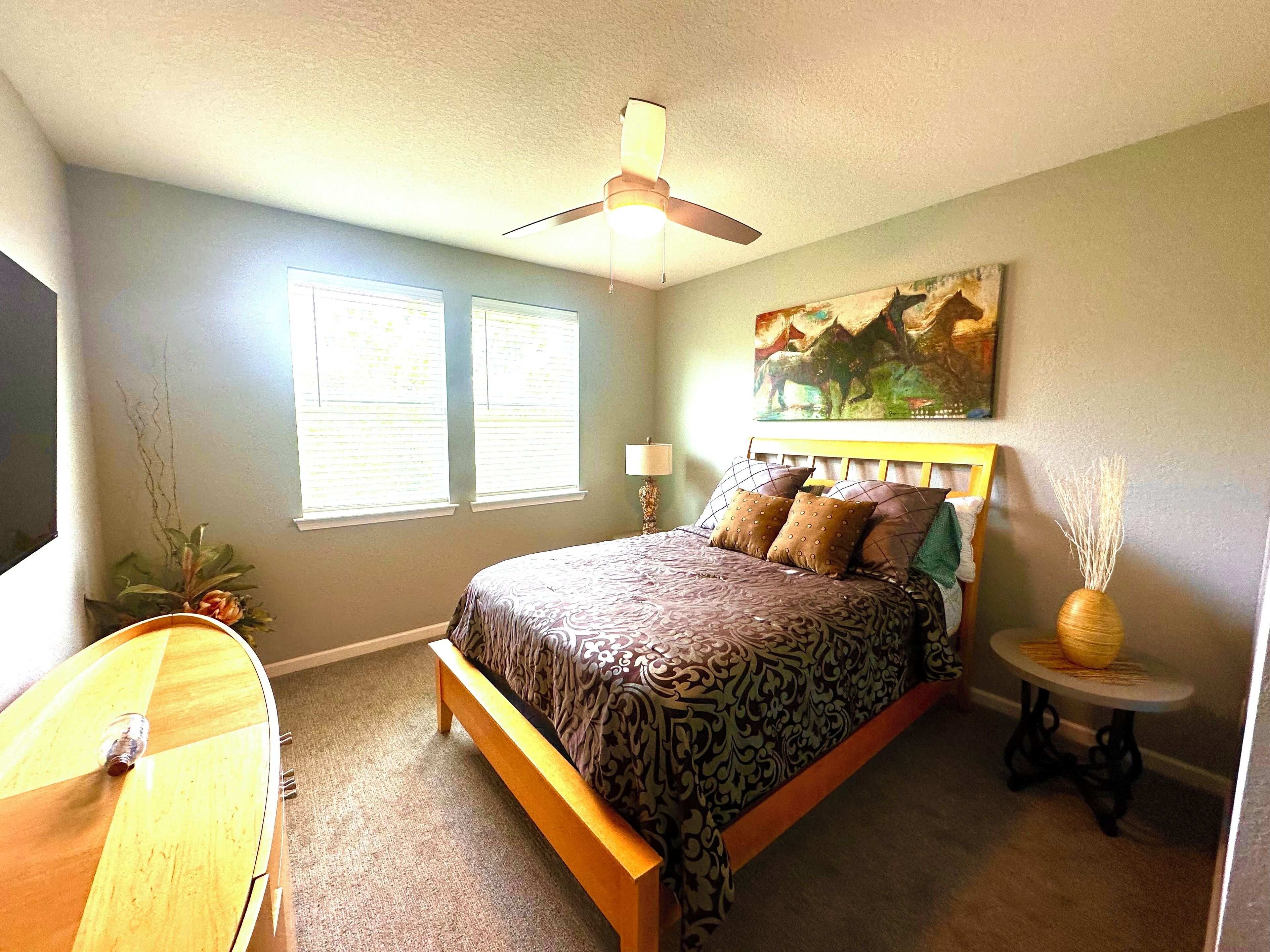 A bedroom with a bed, a ceiling fan, a window with blinds, a painting on the wall, and a small table with a vase.