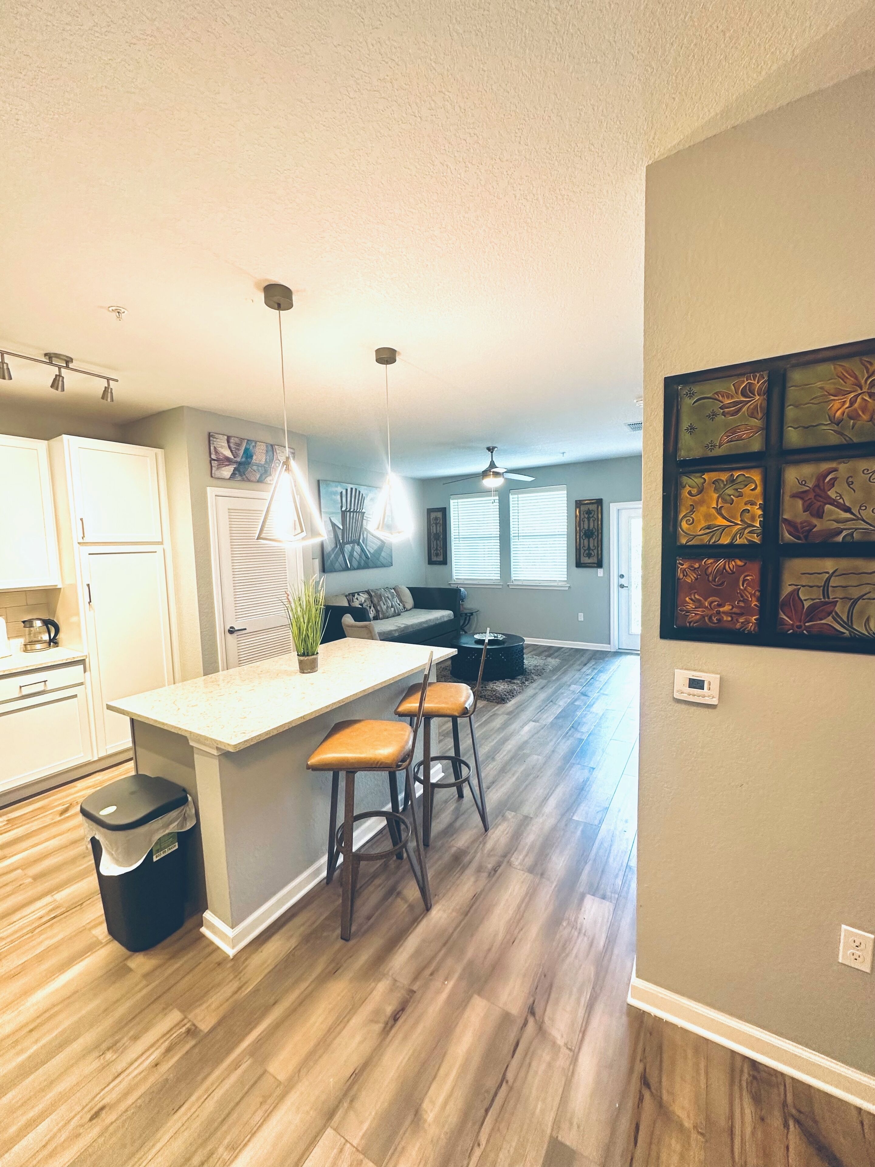 A modern kitchen with a central island, bar stools, pendant lighting, and a dining area with a sofa and coffee table.