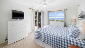 A bedroom with a bed, a TV on a dresser, a ceiling fan, and a view of the sea.
