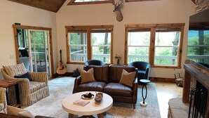 A cozy living room with leather sofas, a coffee table, and a fireplace. Large windows provide natural light and views of the outdoors. - Mountain Views, Hot Tub & Resort Amenities in Sapphire, NC (Sapphire)