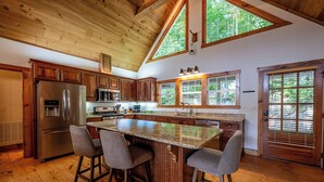 A kitchen with wooden cabinets, a stainless steel refrigerator, a granite countertop, and a large window with a view of trees. - Mountain Views, Hot Tub & Resort Amenities in Sapphire, NC (Sapphire)