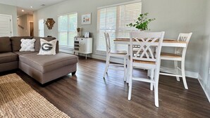 A modern living room with a sectional sofa, a dining table with white chairs, and a small white cabinet. - Chicopee House ~ Your 4 bd, 3 bath, Athens Getaway 1-mile to Sanford Stadium! (Athens)