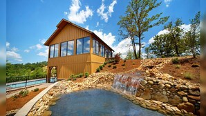 A modern wooden house with large windows, a swimming pool, and a waterfall.