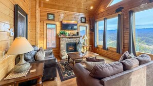 A log cabin interior with a fireplace, leather sofas, a flat-screen TV, and large windows offering a mountain view. - On Eagles Wings・Panoramic Views・Hot Tub・Game Room・Resort Access (Sevierville)