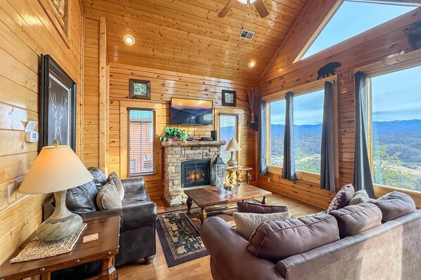 A log cabin interior with a fireplace, leather sofas, a flat-screen TV, and large windows offering a mountain view. - On Eagles Wings・Panoramic Views・Hot Tub・Game Room・Resort Access (Sevierville)