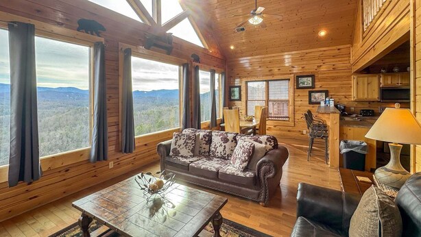 A spacious cabin with a large sofa, dining table, and kitchen area, featuring wooden walls and large windows. - On Eagles Wings・Panoramic Views・Hot Tub・Game Room・Resort Access (Sevierville)