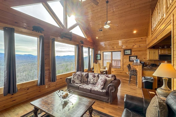 A spacious cabin with a large sofa, dining table, and kitchen area, featuring wooden walls and large windows.