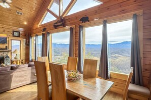 A log cabin interior with a dining area, wooden furniture, large windows, and a fireplace.