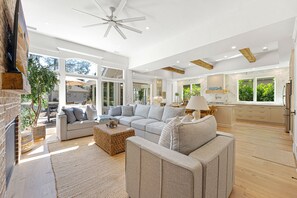 A spacious living area with a large sofa, a coffee table, and a fireplace. There is a kitchen area with cabinets and a window.