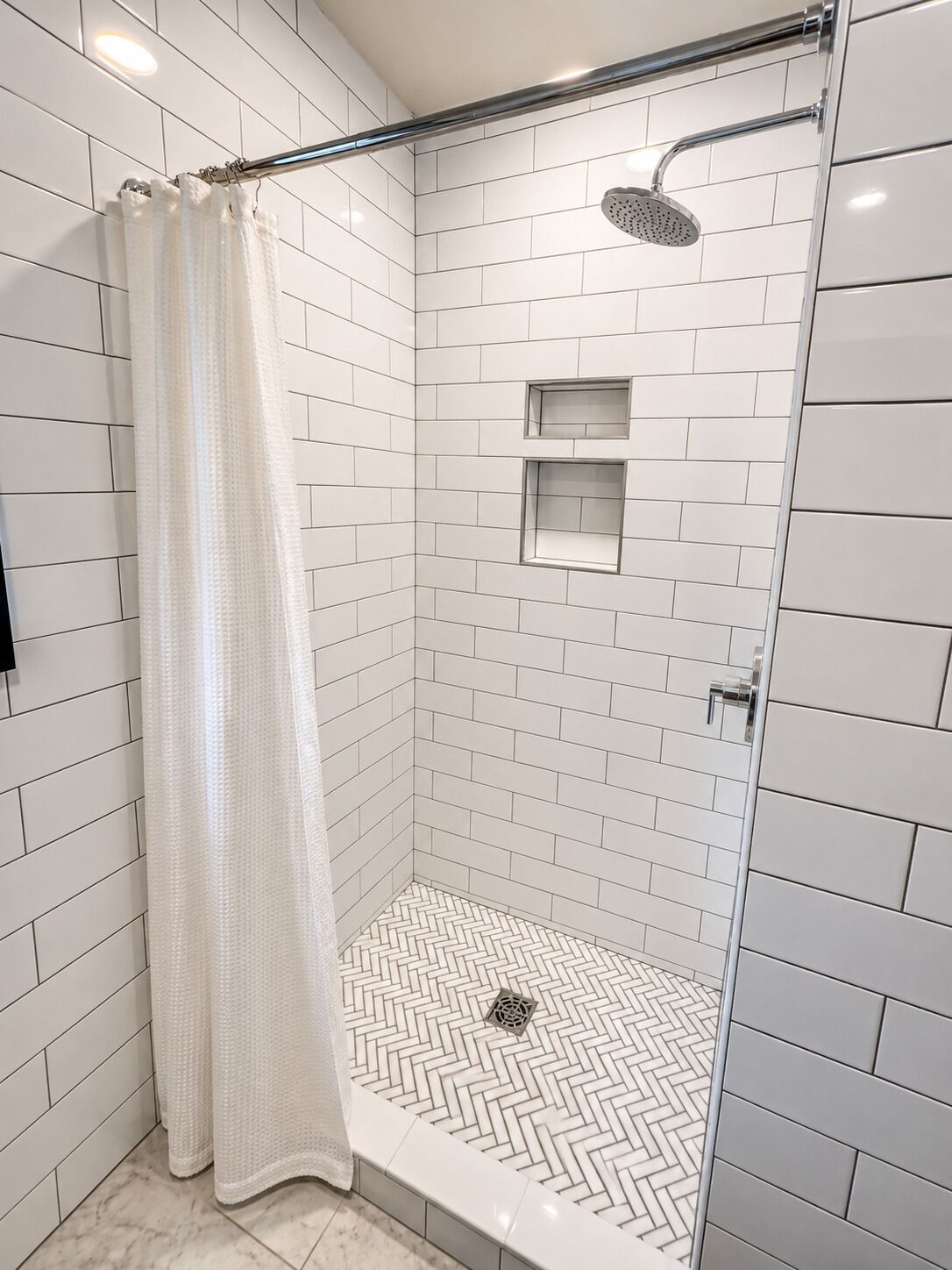 Une douche avec des murs recouverts de carreaux blancs, un sol à motifs en chevrons et un pommeau de douche.