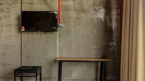 Ένα δωμάτιο με έναν τοίχο από μπετόν, μια τηλεόραση τοποθετημένη στον τοίχο, ένα μικρό μαύρο τραπέζι και ένα ξύλινο τραπέζ ι.