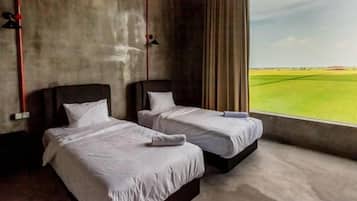 Two beds with white linens in a room with a concrete wall and a large window overlooking a green field.