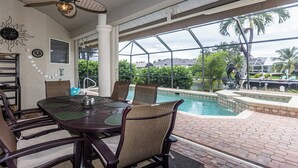 A poolside dining area with a glass-enclosed patio, wicker chairs, and a view of a swimming pool and palm trees.