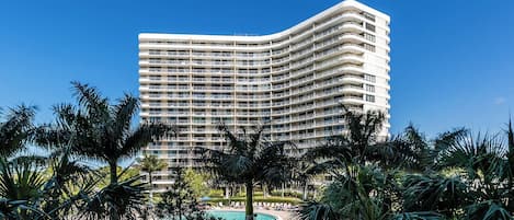 A multi-story hotel with a curved facade, a swimming pool, and palm trees in the foreground.