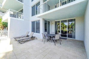 A modern building with a patio area featuring a table and chairs, a sun lounger, and a balcony with railings.