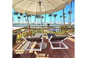 Een houten terras met twee ligstoelen en een parasol, met uitzicht op een zwembadgedeelte met palmbomen en een helder uitzicht op de oceaan.