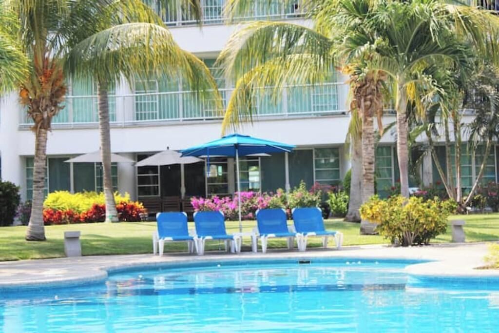 A pool area with blue chairs and umbrellas, surrounded by palm trees and a building with large windows.
