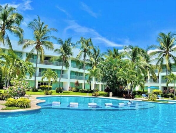 Un resort con una piscina grande, palmeras y un edificio de varios pisos con balcones.