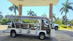 Een witte auto voor drie personen met een dak, geparkeerd onder een constructie met twee grote pilaren en een heldere lucht op de achtergrond.