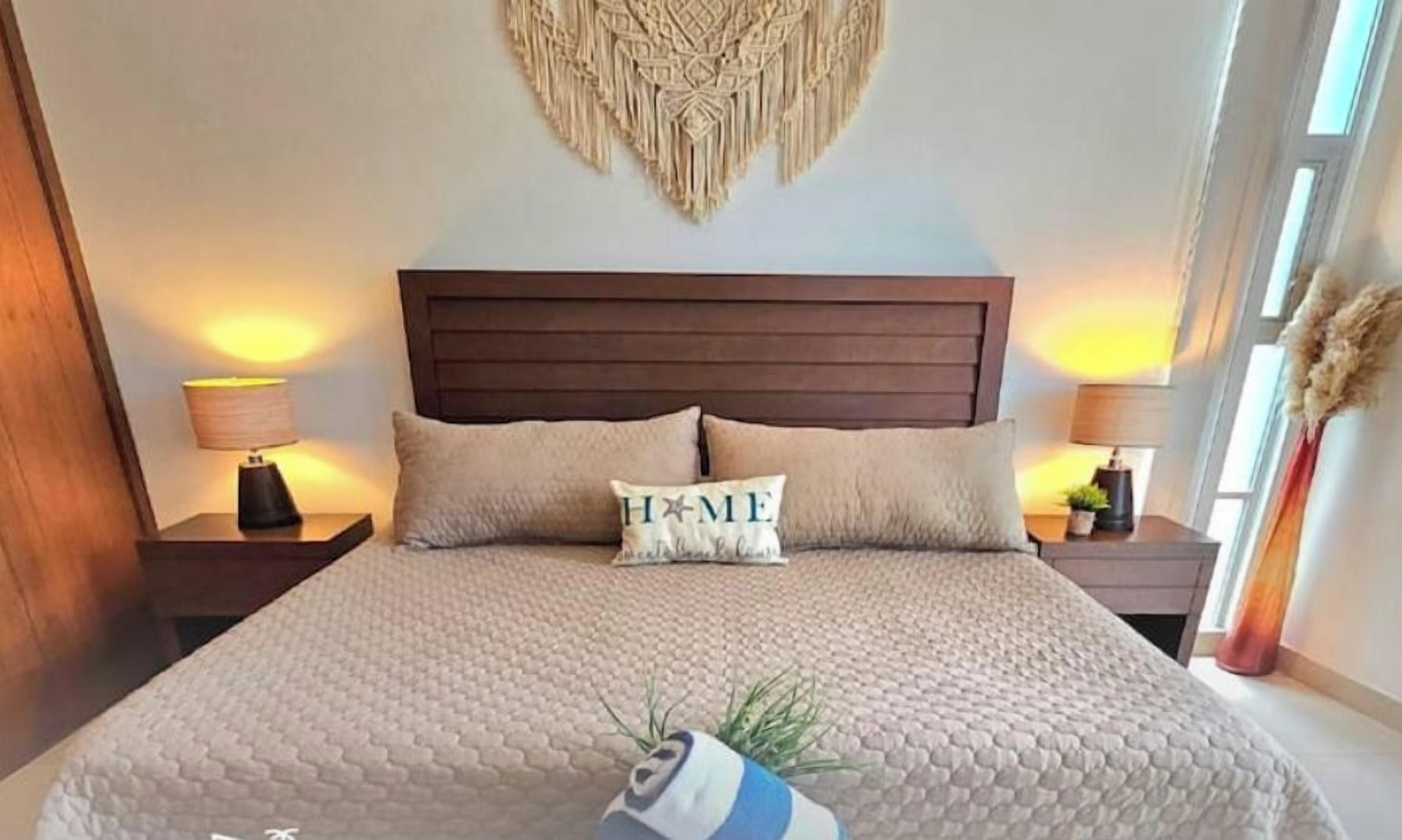 Une chambre avec un grand lit, deux tables de chevet et une décoration murale en macramé.