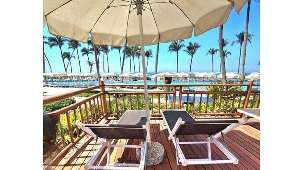 Une terrasse en bois avec deux chaises longues et un parasol, donnant sur une piscine bordée de palmiers et offrant une vue dégagée sur l’océan.
