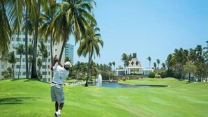 Un terrain de golf où l’on voit une personne vêtue d’un t-shirt et d’un short blanc, prête à frapper une balle de golf, avec des palmiers et un bâtiment en arrière-plan.