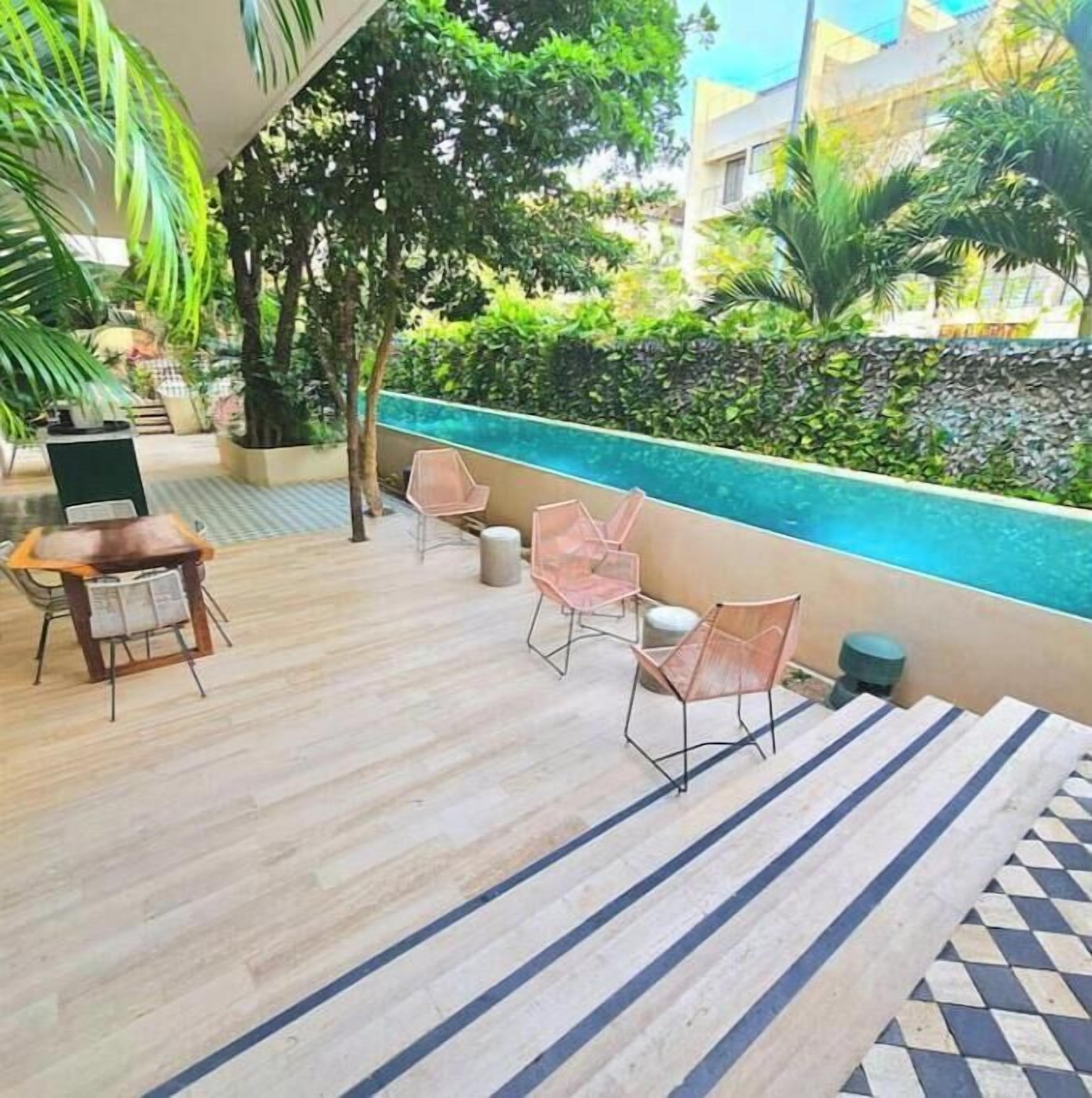 Un espace aménagé au bord de la piscine, avec une table et des chaises en bois, un sol carrelé et une vue sur un bâtiment et de la verdure en arrière-plan.