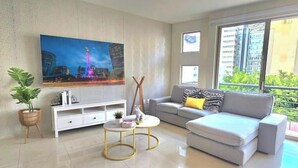 Ein modernes Wohnzimmer mit einem großen Flachbildfernseher, einem grauen Sofa mit einem gemusterten Zierkissen, einem kleinen runden Couchtisch, einer weißen TV-Konsole und einer Topfpflanze.
