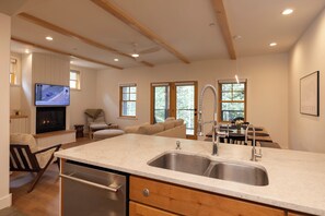 A modern kitchen with a double sink, wooden beams, a dining area with a table and chairs, and a television mounted on the wall.