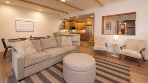A modern living room with a sofa, ottoman, and chairs, a dining area with a table and chairs, and a kitchen with wooden cabinets and appliances.