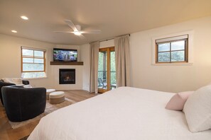 A bedroom with a bed, a television mounted above a fireplace, a ceiling fan, and a balcony with curtains.