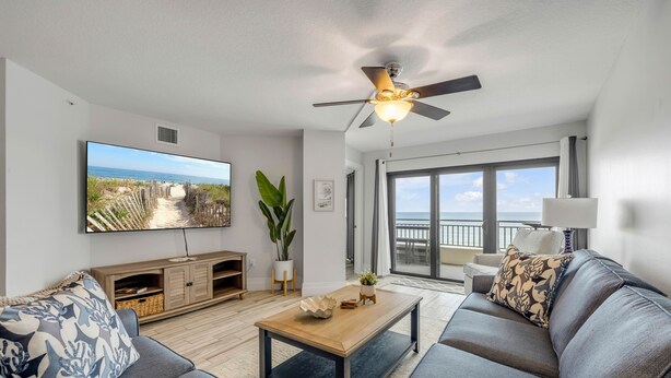 A modern living room with a large flat-screen TV, a ceiling fan, and a view of the ocean through large sliding doors. - New listing! Direct oceanfront 2/2 with private balcony, heated pool, and hot tub (New Smyrna Beach)