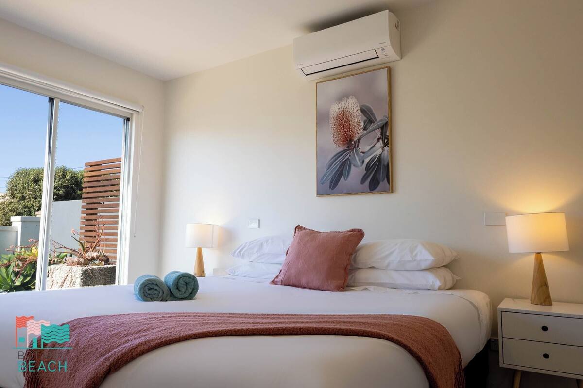 A bedroom with a large bed, two bedside lamps, a wall-mounted air conditioner, and a window with blinds.