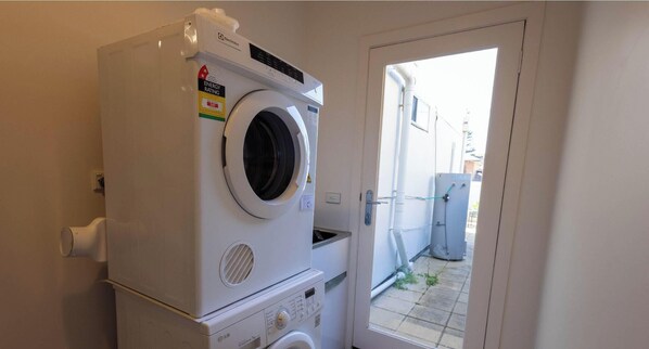 A compact laundry area with a front-loading washer and dryer, a small countertop, and a door leading to an outdoor area. - Botanic Beach (Warrnambool)