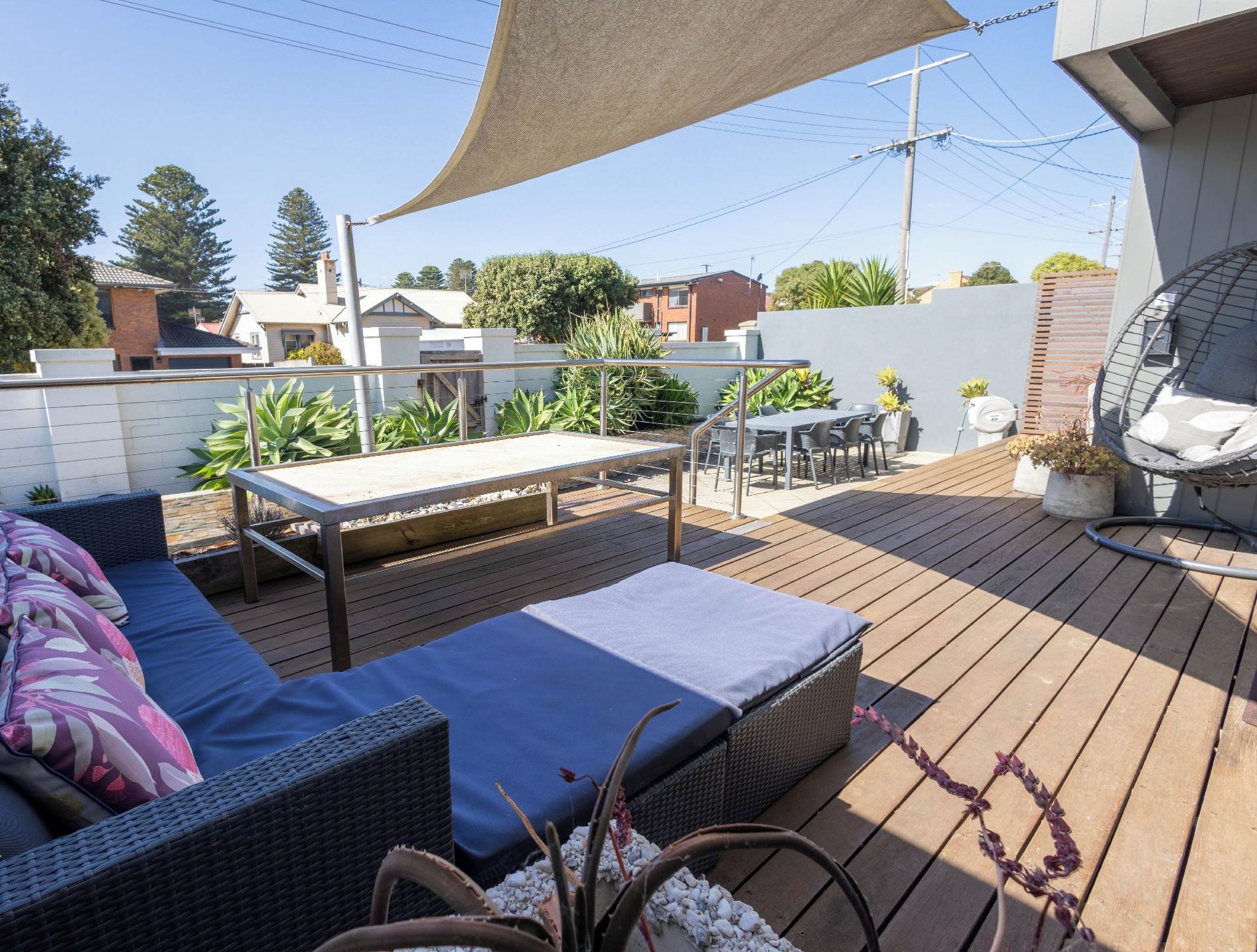An outdoor deck with a sofa, a table, and chairs, surrounded by plants and a shade sail.