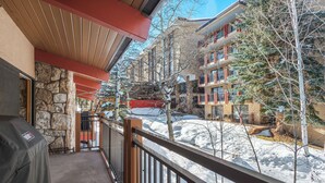 A balcony with a stone wall, a covered area with a red roof, and a view of a snow-covered street with buildings and trees. - Stonebridge Condos Unit 723 (Snowmass Village)