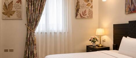 Une chambre d’hôtel avec un lit, une table de chevet avec une lampe, une fenêtre avec des rideaux légers et des œuvres d’art murales.