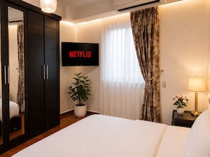 A hotel room with a bed, a television displaying 'NETFLIX,' a potted plant, a nightstand with a lamp, and a mirror.