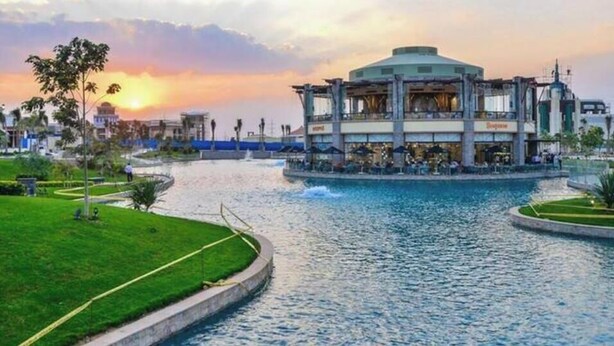 A waterfront hotel with a central building, a swimming pool, and a grassy area.