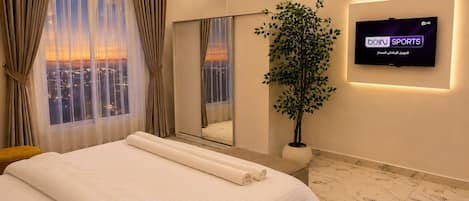 A modern hotel room with a large bed, a television, a potted plant, and a city view.