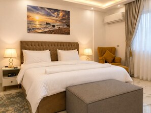 Quarto de hotel com uma cama grande, duas luminárias de cabeceira, uma cadeira, um quadro e um ar condicionado.