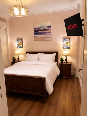 A bedroom with a wooden bed, two bedside tables with lamps, a painting on the wall, and a Netflix sign.
