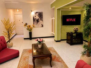 A living room with a sofa, a coffee table, a painting, a TV displaying 'NETFLIX', and a potted plant.