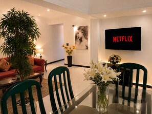 Ein modernes Wohnzimmer mit einer Couch, Sesseln, einem Esstisch, einem Topfpflanzen und einem an der Wand befestigten Fernseher, der 'NETFLIX' anzeigt.