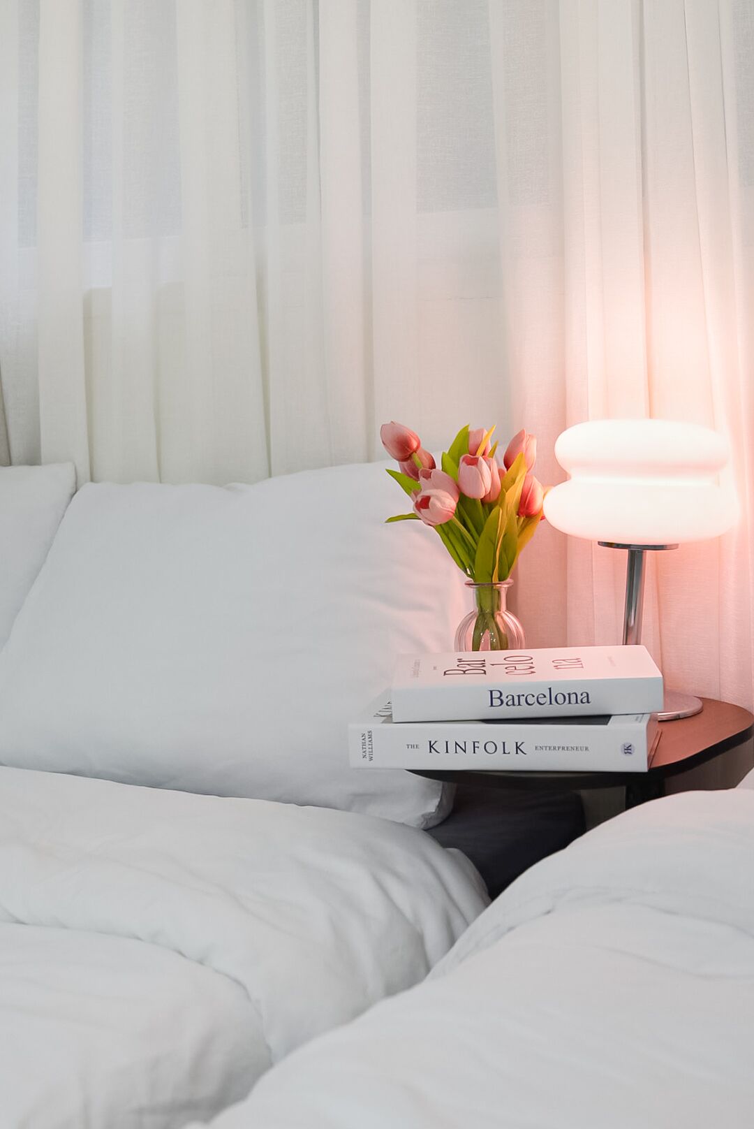 A neatly made bed with white bedding, a bedside table with a lamp, a vase with pink tulips, and a stack of books.