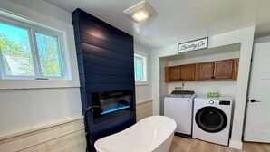 A compact laundry room with a built-in washer and dryer, a white freestanding bathtub, and wooden cabinets above the appliances. - 91M: Renovated Bethlehem home with hot tub, pool, firepit, fenced yard! Walk to restaurants (Bethlehem)