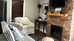 Ett vardagsrum med en grå soffa, en rund pall, en öppen spis, en väggmonterad TV och en takfläkt.