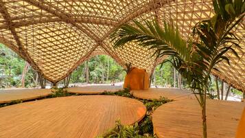 Un pabellón descubierto con una estructura de techo de bambú geométrica, rodeado de exuberante vegetación y pasarelas de madera.