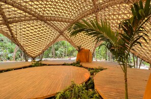 Un pavillon à ciel ouvert doté d’une structure de toit géométrique en bambou, entouré d’une végétation luxuriante et de passerelles en bois.
